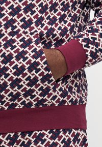 Mønstret pullover i kastanje, elfenben og marineblå med et geometrisk design. Manchetter og talje i ensfarvet kastanje. Sidelommen er synlig.