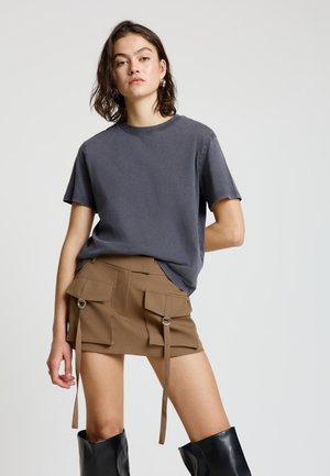 Giovane donna che indossa una maglietta grigio scuro, una minigonna marrone con grandi tasche e cinturini, e stivali neri alti fino al ginocchio, in piedi davanti a uno sfondo semplice.