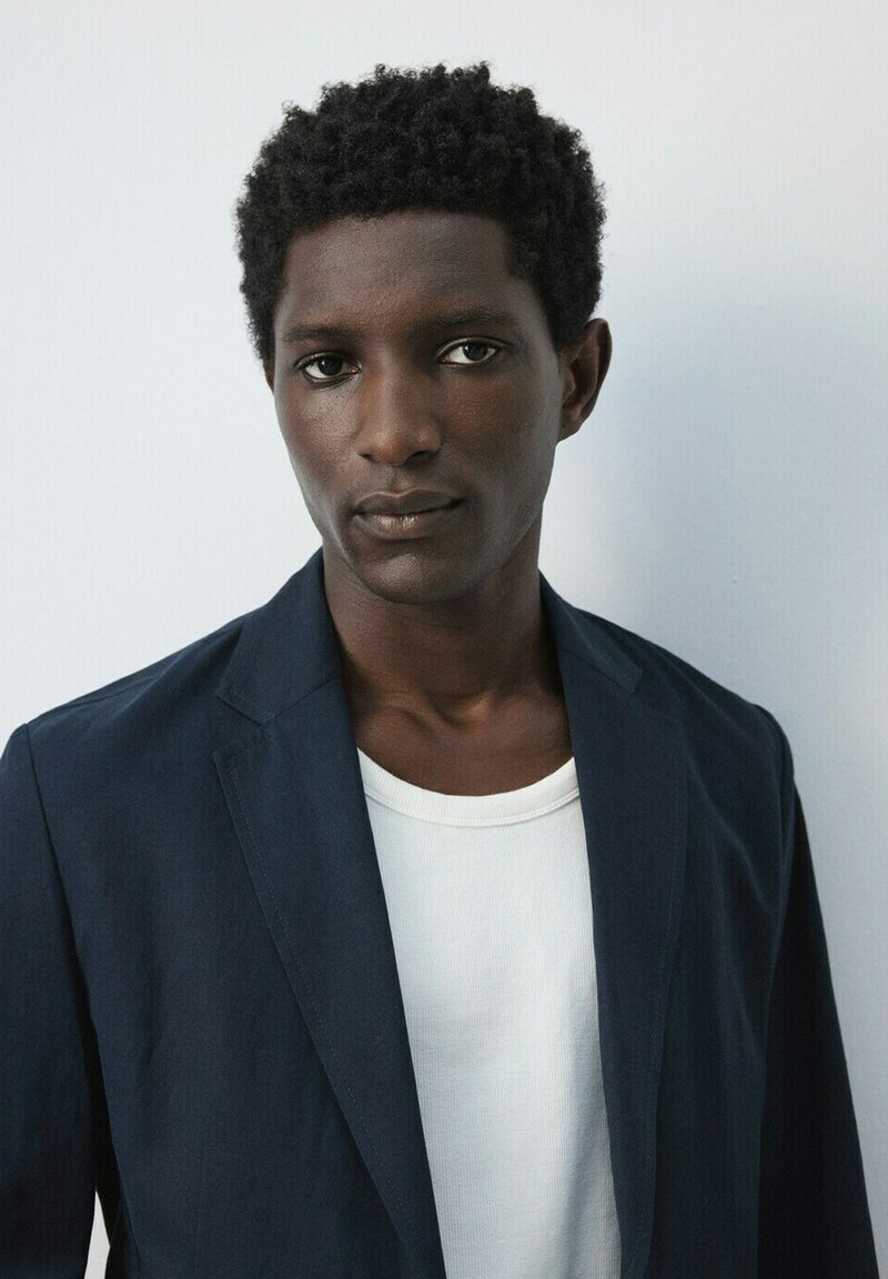 Joven con cabello corto y rizado, vistiendo un blazer oscuro sobre una camisa blanca, de pie frente a un fondo liso de color gris claro.