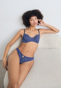Blue lace bra and matching brief set featuring intricate floral patterns, satin accents, and adjustable straps. Model seated on textured surface.