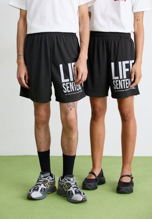 Schwarze Netz-Trainingsshorts mit dem weißen Aufdruck "LIFE SENTENCED". Kombiniert mit grauen Sneakers und schwarzem Schuhwerk. Einfarbige weiße T-Shirts.