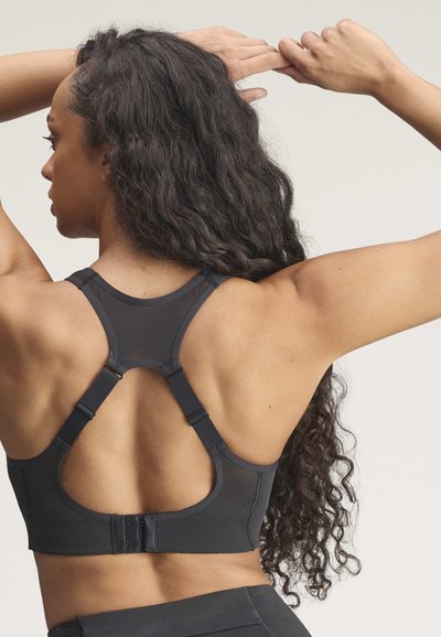 Kvinde med langt krøllet hår iført en sort sports-bh med justerbare stropper og lukning med krog og øje på ryggen, løfter armene bag hovedet.