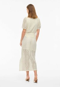 Robe midi en crochet crème avec des manches courtes bouffantes, des motifs floraux, une taille cintrée et une fente sur le côté, associée à des sandales à talons beiges.