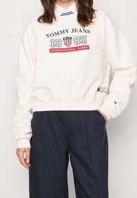 Kremowa bluza z okrągłym dekoltem, z logo "TOMMY JEANS 1985 INTERNATIONAL GAMES" w granatowym i czerwonym kolorze. Nieco skrócony krój.