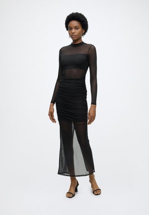 Kobieta stojąca w długiej czarnej prześwitującej sukience z marszczonymi detalami, długimi rękawami i czarnymi sandałami na wysokim obcasie na jednolitym białym tle.