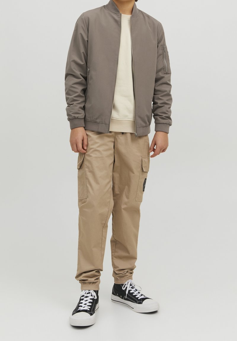 Niño vestido con pantalones cargo beige, una chaqueta bomber topo, un sudadera de color crema y zapatillas altas negras sobre un fondo simple.
