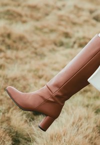 Cizma din piele maro, până la genunchi, cu vârful rotund și toc gros. Prezintă o textură netedă și un design simplu, cu cusături verticale.