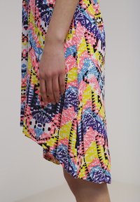 Main reposée contre une jupe de longueur genou avec des motifs géométriques colorés en rose, jaune, bleu et noir sur un fond clair.