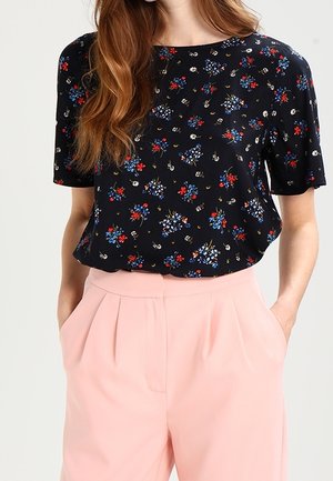 Nainen pukeutuneena mustaan kukkakuosiseen lyhythihaiseen puseroon, joka on pantu korkeavyötäröisten, laskostettujen vaaleanpunaisten housujen sisään, kädet taskuissa.