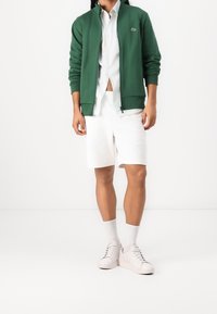 Sudadera verde con cremallera, de mezcla de algodón, combinada con una camisa blanca abotonada y pantalones cortos blancos. Zapatillas blancas con cordones y calcetines a media pierna.
