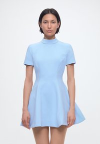 Γυναίκα με κοντά σκούρα μαλλιά που φοράει εφαρμοστό ανοιχτό μπλε μίνι φόρεμα με κοντά μανίκια και υψηλό λαιμόκοψη, σε απλό λευκό φόντο.