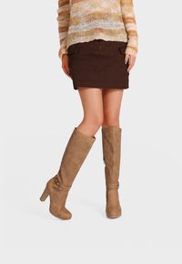 Mujer con minifalda marrón, suéter de punto beige y botas perforadas de tacón color cuero hasta la rodilla, de pie con las piernas cruzadas.