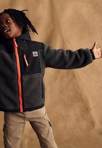 Child with dreadlocks wearing a black and gray fleece jacket with orange zippers and beige cargo pants, extending one arm outward.