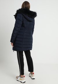 Mörkblå puffjacka med huva av konstgjord päls, elastisk midja, långa ärmar och quiltat mönster; matchad med svarta träningsbyxor och sneakers.