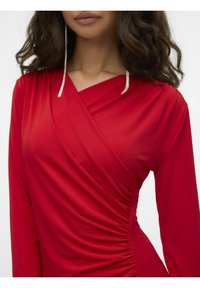 Top rosso avvolgente in tessuto morbido con maniche lunghe, dettaglio arricciato sui lati e scollatura a V profonda, completato da orecchini d'argento.