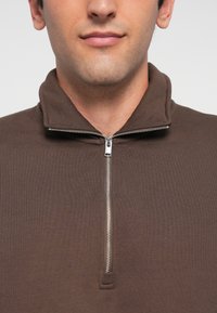 Ung man som bär en brun tröja med kvartdragkedja där dragkedjan är delvis stängd, vilket visar kragen och nedre delen av ansiktet.