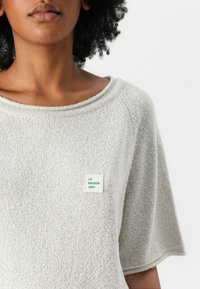 Light gray, textured sweater with a wide neckline and short sleeves. Features a square label with "LA MAISON AMV" in green.