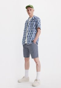Camisa de cuadros azul y blanca de manga corta, pantalones cortos grises, gorra verde, zapatos claros. El modelo está de pie con las manos en los bolsillos frente a un fondo blanco.