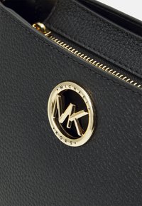 Bolso de piel negra texturizada que presenta un logo circular dorado con el texto "Michael Kors" y las iniciales dobles "MK", acentuado con una cremallera dorada.