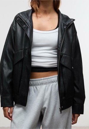 Personne portant une veste en cuir noire par-dessus un crop top blanc et un pantalon de survêtement gris clair, présentée des épaules aux hanches sur un fond uni.