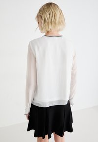 Haut en chiffon blanc avec des manches longues transparentes, accents noirs, et une jupe noire unie avec un ourlet évasé. Aucuns motifs ni accessoires visibles.