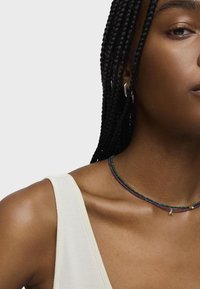Collana choker multicolore con perline verdi, viola e blu. Presenta un piccolo charm e viene indossata con una canotta bianca a coste.