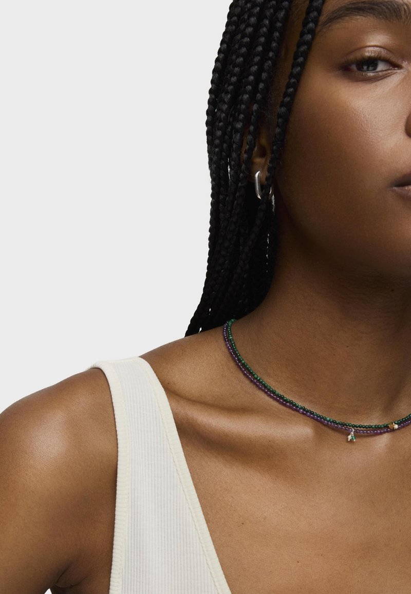 Collana choker multicolore con perline verdi, viola e blu. Presenta un piccolo charm e viene indossata con una canotta bianca a coste.