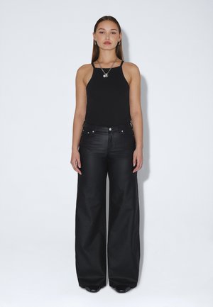 Woman standing against a plain background wearing a black sleeveless top, wide-leg black pants, black shoes, and silver necklaces.