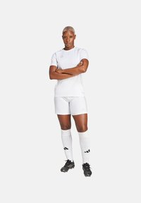 Wit sportshirt en short met subtiele textuur, gecombineerd met knielange witte sokken en zwarte voetbalschoenen. Bevat Adidas-branding.