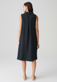 Sleeveless navy dress with a collar, featuring a pleat detail at the back. Made from a smooth, lightweight fabric. Mid-length design.