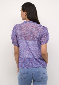 Blusa viola trasparente con motivo floreale, maniche a sbuffo corte e scollatura arricciata. Abbinata a jeans blu aderenti con tasche posteriori.