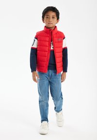 Röd pufferväst med hög krage, bärs över en marinblå tröja med vita och röda ränder, kombinerat med ljusblå jeans och vita sneakers.