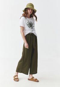 White t-shirt with green leaf print, olive-green wide-leg pants with a tied waist, and tan slide sandals. Green crocheted bucket hat.