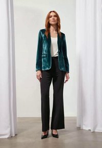 Velvetové sako s klopami v tyrkysovej farbe, zapínanie na jeden gombík a dva predné vrecká, v kombinácii s čiernymi rozšírenými nohavicami a čiernymi vysokými opätkami.
