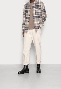 Man in een beige en witte geruite overhemd, taupe t-shirt, off-white broek opgerold aan de mouwen, en zwarte leren enkelboots op een grijze vloer.