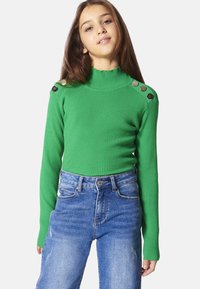 Maglione verde a coste con colletto alto e maniche lunghe, dotato di bottoni metallici sulle spalle. Abbinato a jeans in denim blu a vita alta.