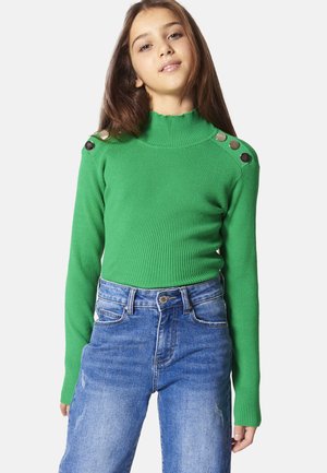 Maglione verde a coste con colletto alto e maniche lunghe, dotato di bottoni metallici sulle spalle. Abbinato a jeans in denim blu a vita alta.