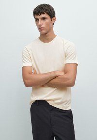 T-shirt crema a maniche corte con scollo rotondo, texture morbida e vestibilità rilassata, abbinato a pantaloni scuri. Design semplice e minimalista.