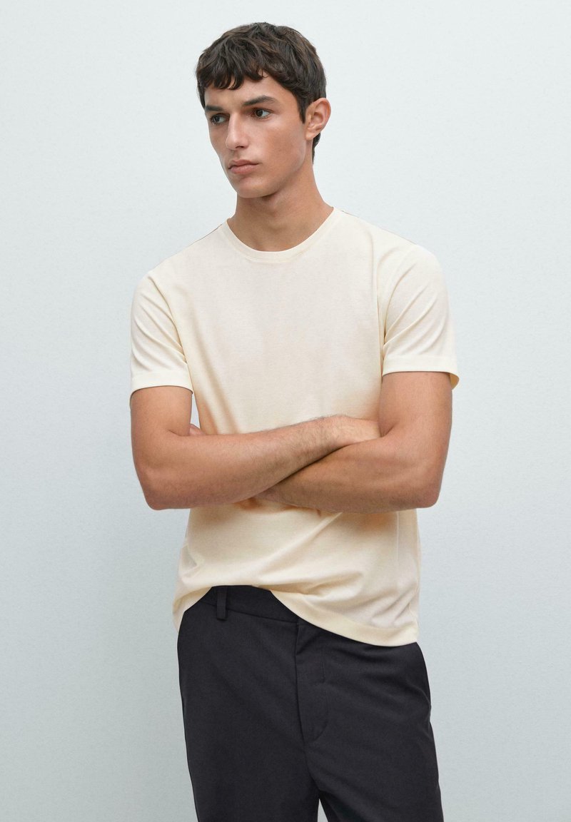 T-shirt crema a maniche corte con scollo rotondo, texture morbida e vestibilità rilassata, abbinato a pantaloni scuri. Design semplice e minimalista.