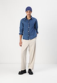 Camicia button-up in denim blu, con colletto classico e maniche arrotolate. Pantaloni beige larghi e scarpe nere robuste.