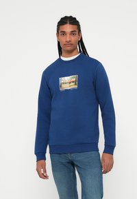 Sudadera azul en mezcla de algodón con cuello redondo, con un gráfico central de un diner. Llevada con jeans azul claro, el modelo está de pie contra un fondo blanco.
