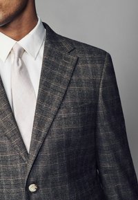 Grau gemusterte Anzugjacke mit Karo-Muster, mit Reverskragen, einer Ein-Knopf-Verschluss und einer hellen Krawatte über einem weißen Hemd.