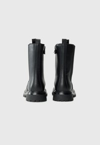 Une paire de bottes en cuir noires mi-mollet avec des semelles chunky et des tirants, vues de l'arrière sur un fond clair et uni.