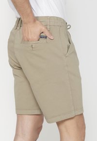 Vīrietis, kurš valkā bēšus šortus ar roku aizmugurē esošajā kabatā, uz kuras redzams melns Koroshi uzlīme, pret vienkāršu baltu fonu.