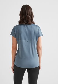 Woman with shoulder-length brown hair wearing a blue short-sleeve top and black leggings, viewed from the back against a plain background.