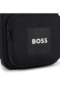 Η μαύρη τσάντα από νάιλον διαθέτει λεία υφή, φερμουάρ κλεισίματος και ένα ορθογώνιο τσεπάκι με το λευκό λογότυπο "BOSS" σε μαύρο φόντο.