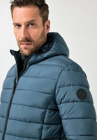 Giacca imbottita in turchese, con chiusura a zip, cappuccio e dettagli a coste. Tessuto strutturato con cuciture e logo circolare sulla manica.