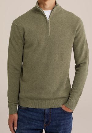 Mann trägt einen olivgrünen, strukturierten Pullover mit kurzem Reißverschluss über einem weißen Hemd und blauen Jeans, eine Hand in der Tasche.