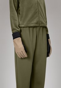 Veste zippée en polaire olive, associée à un pantalon olivier ample, avec des gants beige et noir sur la main droite.