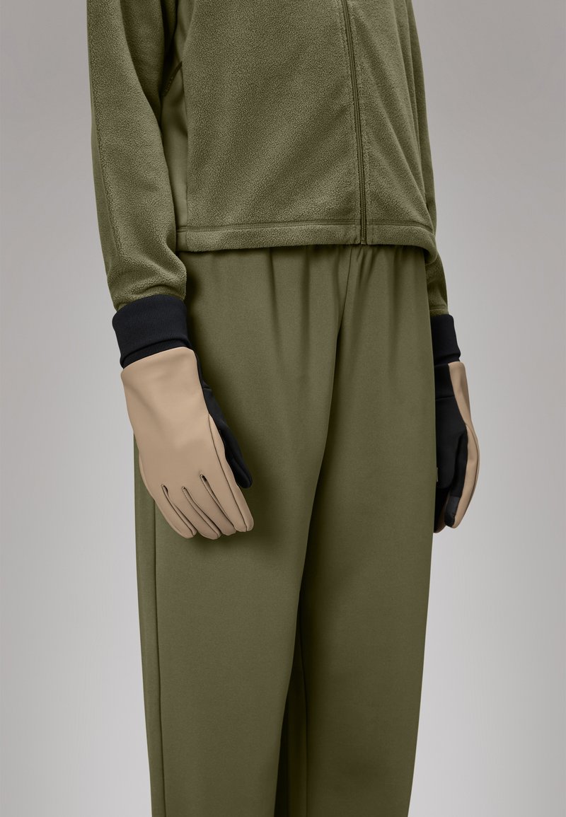 Veste zippée en polaire olive, associée à un pantalon olivier ample, avec des gants beige et noir sur la main droite.